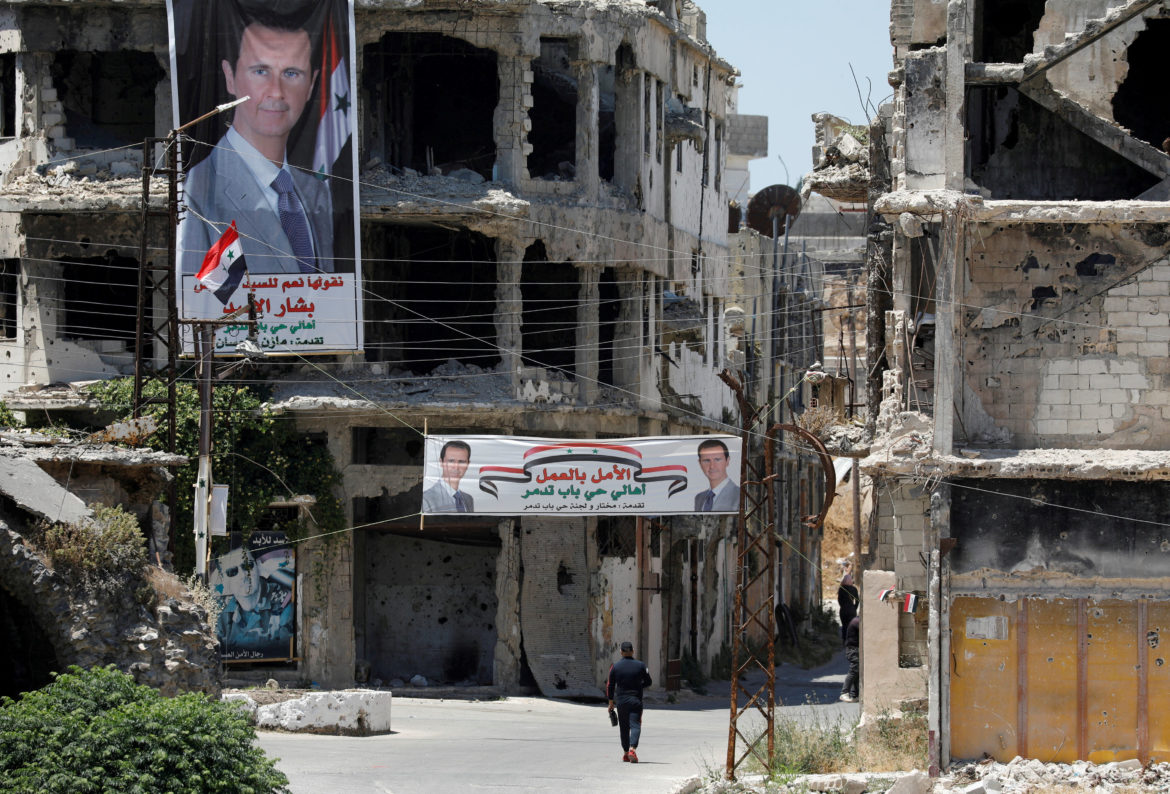 FILE PHOTO: A man walks past banners depiciting Syria’s President Bashar al-Assad, near damaged buildings in Homs